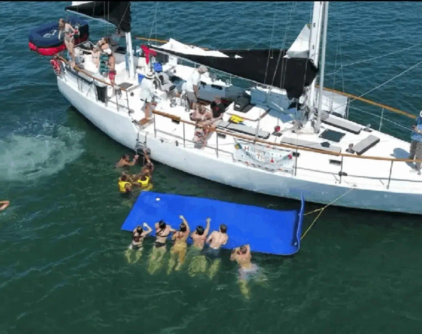 People swimming and relaxing next to a sailboat on the water.