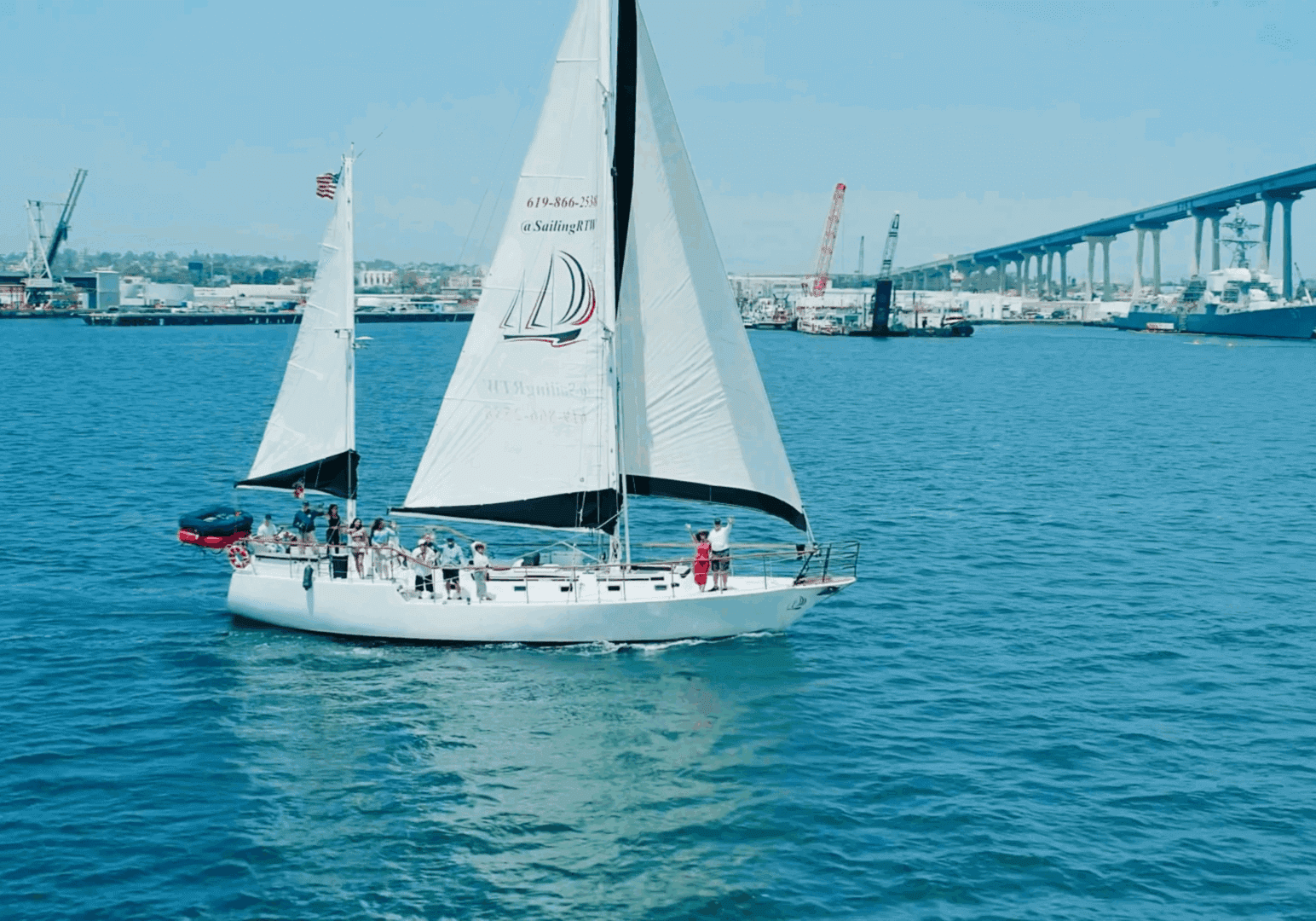 Sailboat glides on calm blue waters near a city harbor.