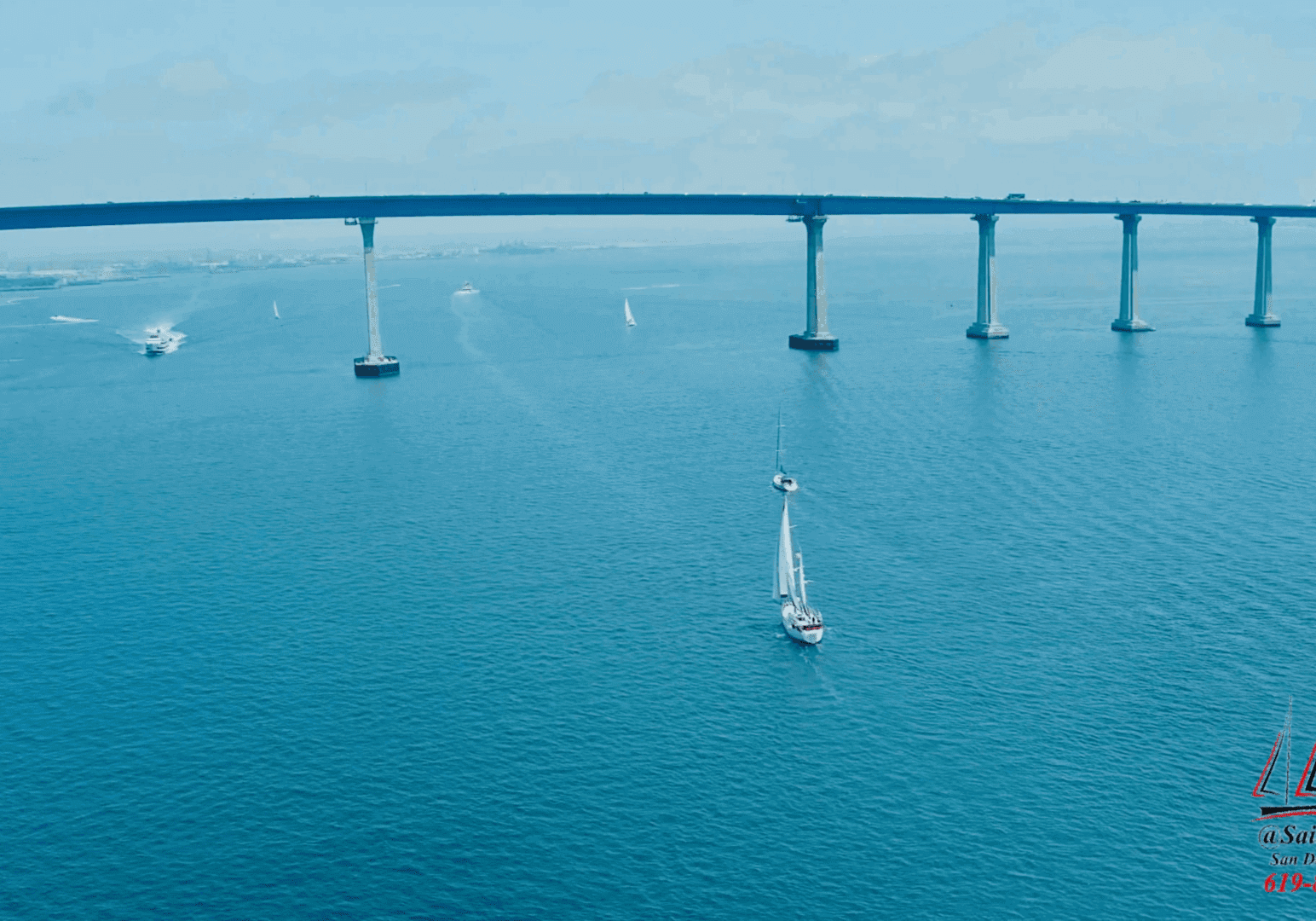 A sailboat glides under a long bridge over clear blue water.