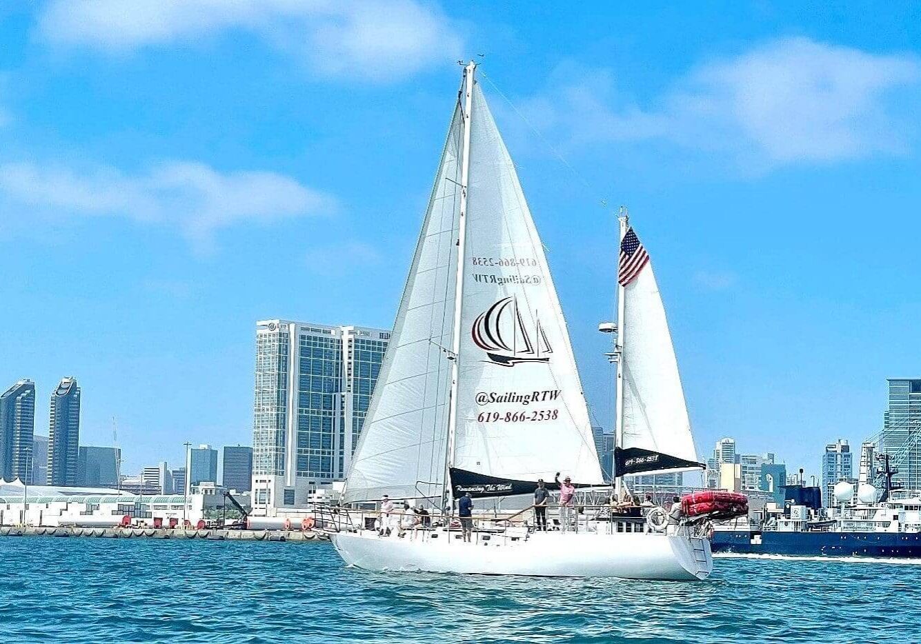 A sailboat with white sails cruising near a city skyline under a blue sky.